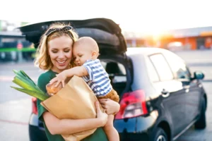 A smiling parent holds a baby while unloading groceries from a car in a parking lot.