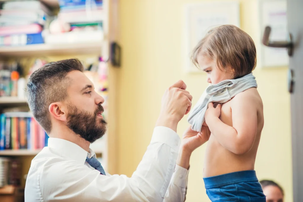 An adult helps a toddler pull on a shirt while standing indoors during a caregiving routine.