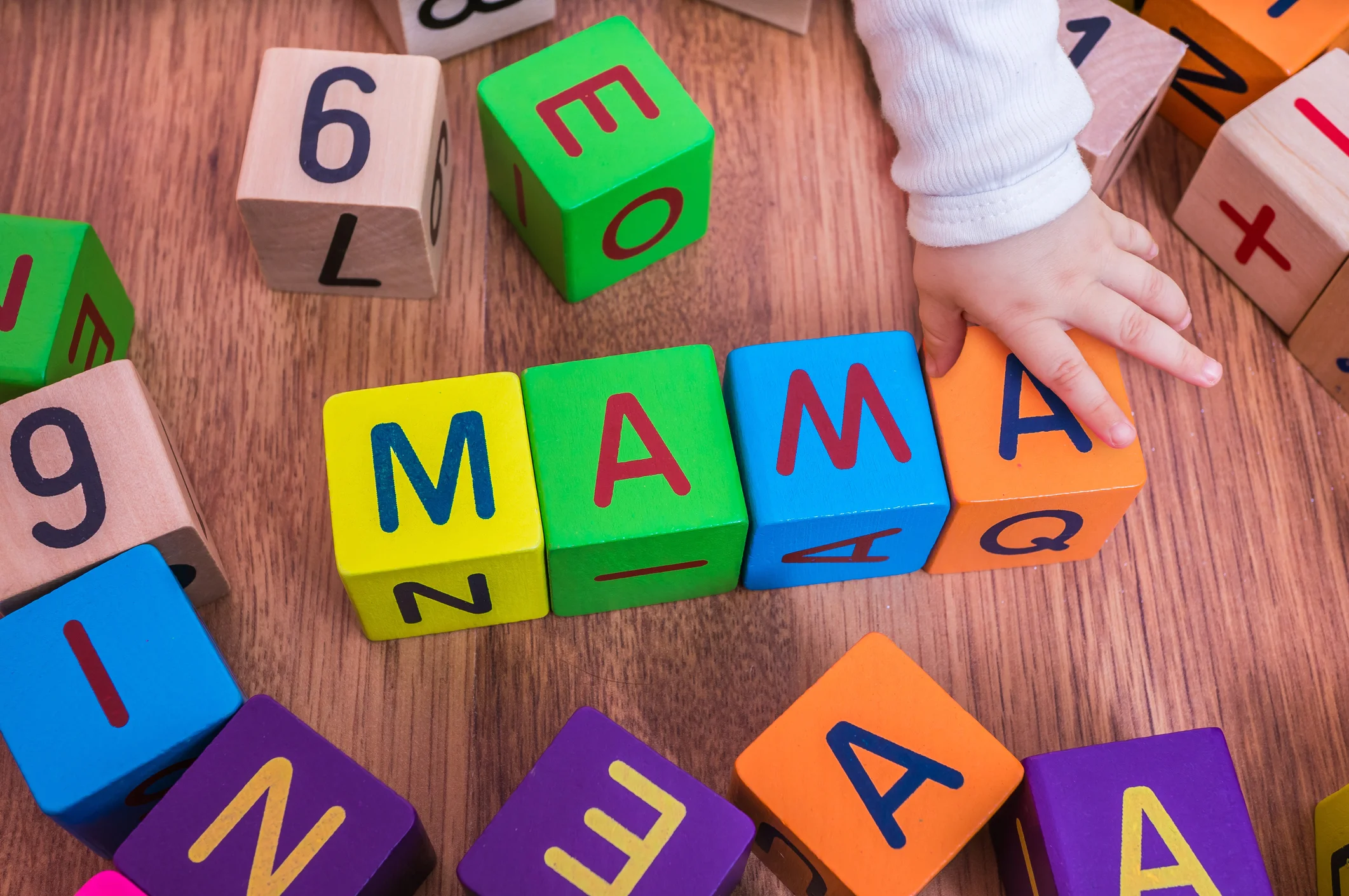 A child’s hand arranges colorful wooden letter blocks spelling “MAMA” on a wooden floor.