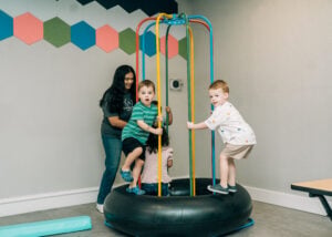 Children stand and sit on a spinning therapy platform while a therapist supports balance and movement.