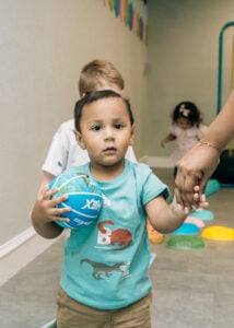 A toddler walks forward holding a small ball while an adult provides hand support during therapy.