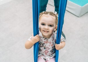 A smiling child holds onto a fabric swing while seated and supported during a sensory activity.