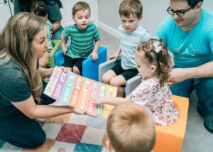 A therapist leads a small group of children during circle time using a colorful visual chart.
