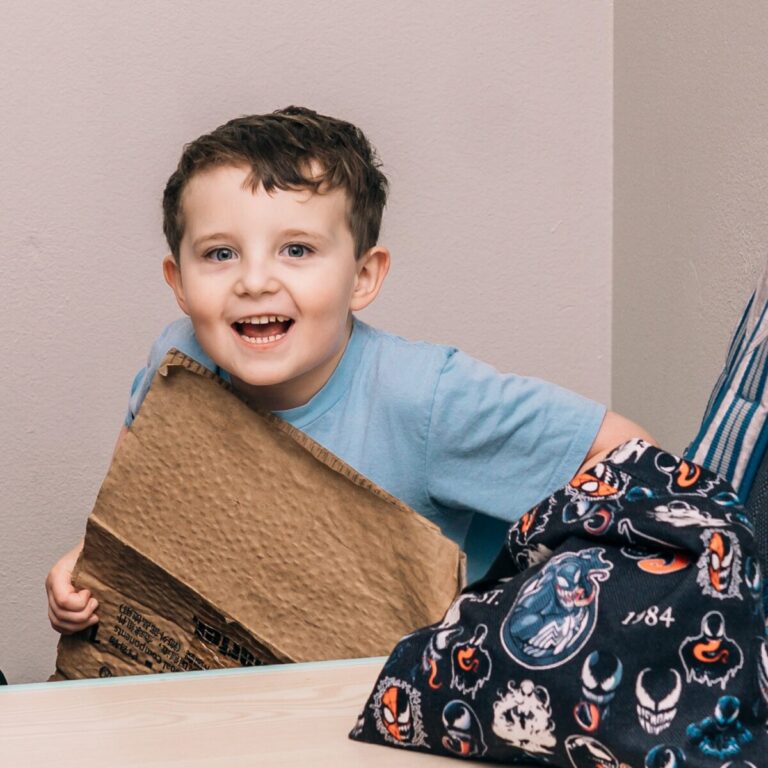 A happy child sits at a table holding a textured bag during a hands-on sensory activity.