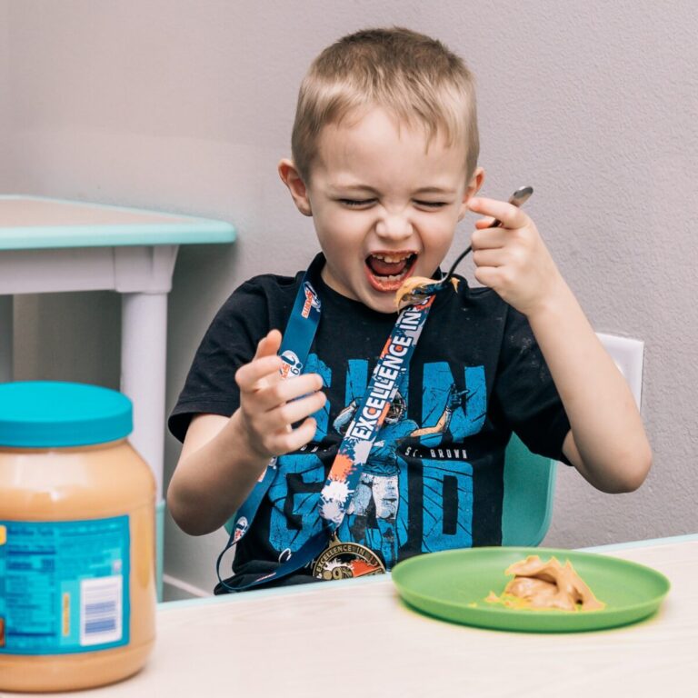 A smiling child eats from a spoon at a table with a plate of food and a jar nearby.