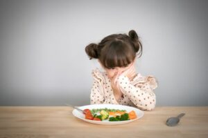 A young child sits at a table covering their mouth while looking at a plate of vegetables.