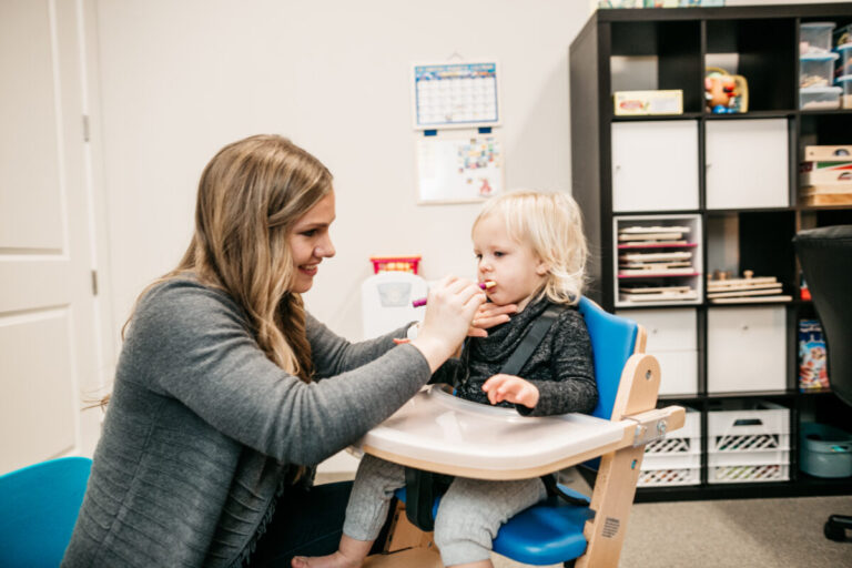 A therapist gently feeds a toddler seated in a high chair during a guided feeding activity.